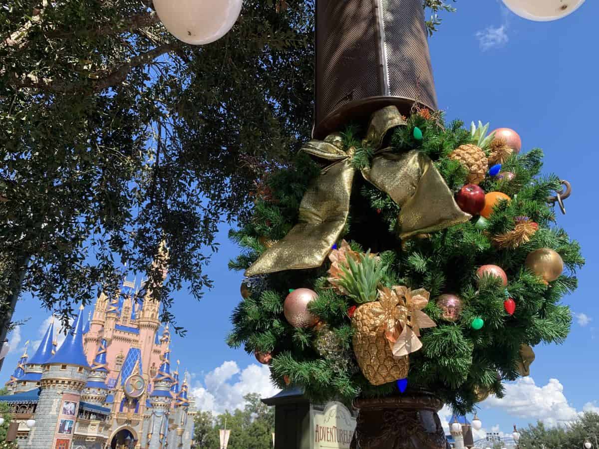A festive wreath adorned with a gold bow and pineapples decorates a lamppost on Main Street, U.S.A. near Cinderella Castle at Magic Kingdom, set against a bright blue sky. The scene captures the enchanting holiday spirit of Disney Very Merriest After Hours, the exclusive sold-out event lighting up special nights at Walt Disney World.