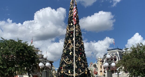 The grand Christmas tree at Magic Kingdom, decked out in festive ornaments with an American flag at its base, stands tall in the heart of Main Street, U.S.A. Guests stroll around, taking in iconic Disney buildings and the holiday magic under a vibrant blue sky—a classic Disney World holiday celebration.