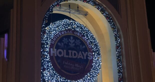 A dazzling holiday wreath illuminated with twinkling lights and a "Universal Orlando Holidays" sign welcomes guests at the arched entrance to Universal Orlando Resort, creating festive nighttime magic.