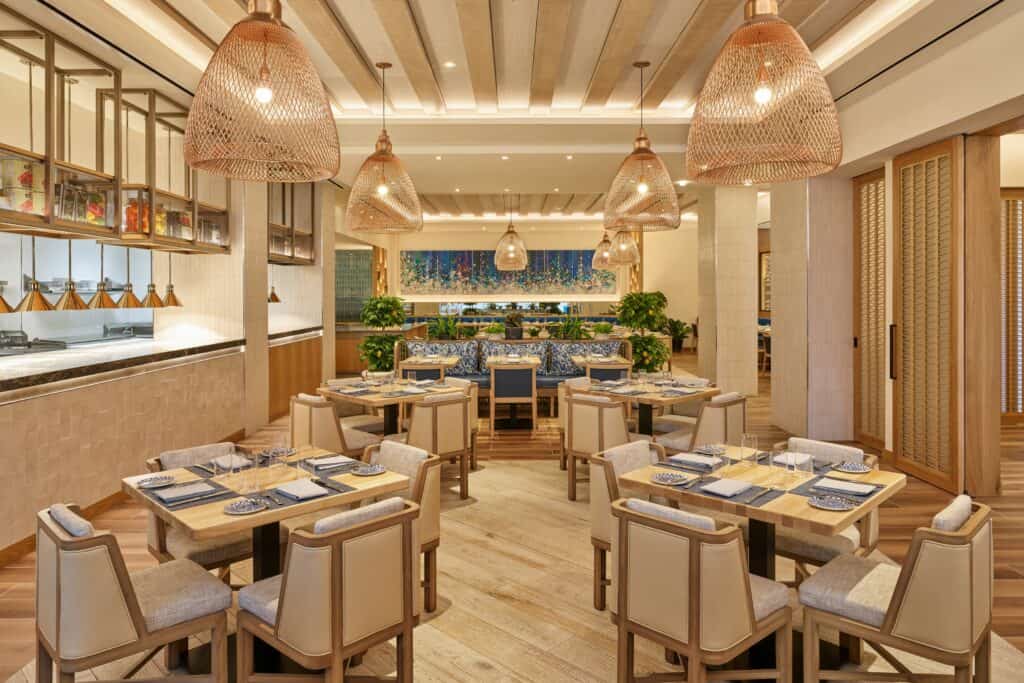 Chic dining space at the Swan Reserve in Walt Disney World, featuring stylish wooden tables, cream-colored chairs, glowing hanging lights, and vibrant blue touches—a modern spot for resort guests to unwind after a day in the parks.