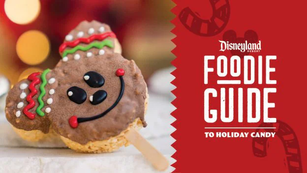 Festive Mickey-shaped treats on sticks displayed beside the vibrant red Disneyland Foodie Guide to Holiday Candy sign, offering fans a magical way to enjoy seasonal sweets during their park adventure.