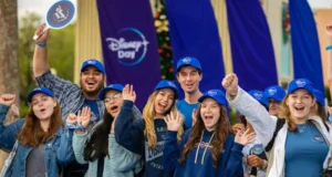 Disney fans decked out in blue hats and Disney shirts gather outdoors at a Disney Parks celebration, beaming with excitement as they cheer for a truly Blue-tiful Day.