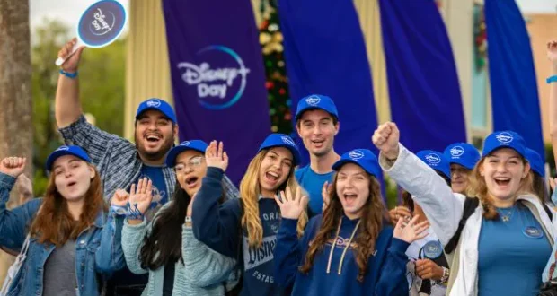 Disney fans decked out in blue hats and Disney shirts gather outdoors at a Disney Parks celebration, beaming with excitement as they cheer for a truly Blue-tiful Day.