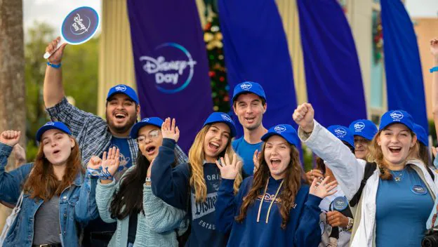 Disney fans decked out in blue hats and Disney shirts gather outdoors at a Disney Parks celebration, beaming with excitement as they cheer for a truly Blue-tiful Day.