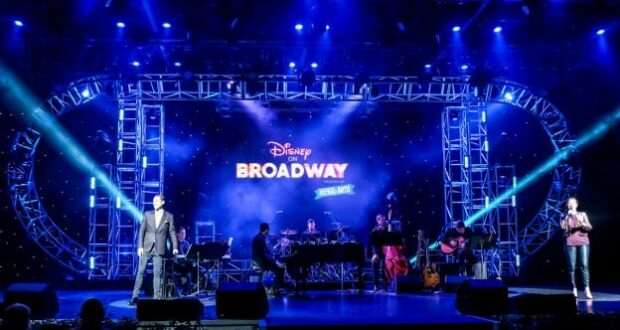 Talented performers sing and play music live on the EPCOT stage during the Disney on Broadway concert series, with the iconic "Disney on Broadway" logo glowing brightly on a giant screen behind them.