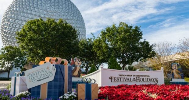 A festive display at Epcot features large wrapped gifts and a sign reading "Epcot International Festival of the Holidays." Colorful flowers and two cartoon character cutouts are in the foreground, with Spaceship Earth in the background.