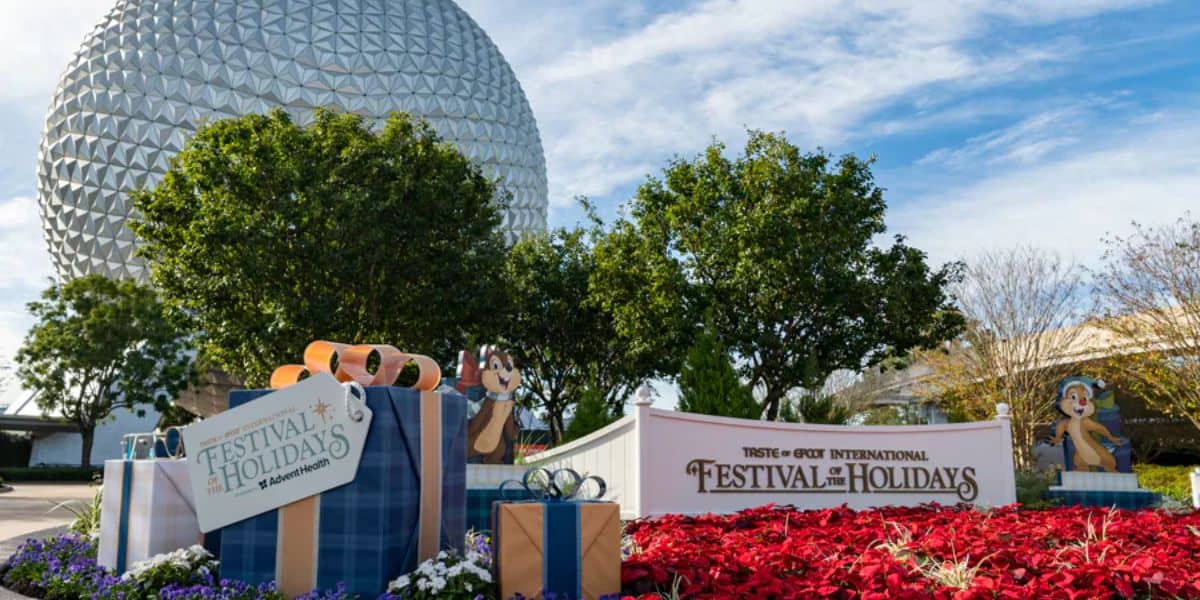 A festive display at Epcot features large wrapped gifts and a sign reading "Epcot International Festival of the Holidays." Colorful flowers and two cartoon character cutouts are in the foreground, with Spaceship Earth in the background.