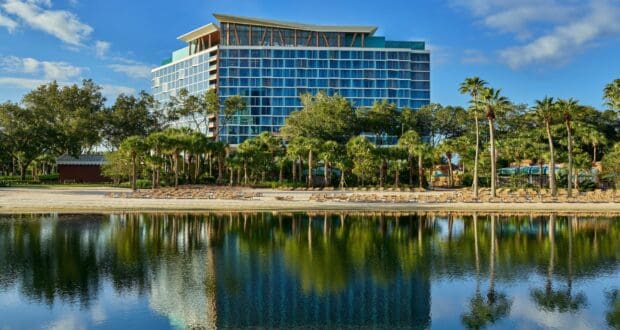 The Swan Reserve Hotel at Walt Disney World features striking blue windows, swaying palm trees, and a shimmering lake reflecting the vibrant sky—offering guests an elegant and immersive resort experience just steps from the magic of Disney’s theme parks.