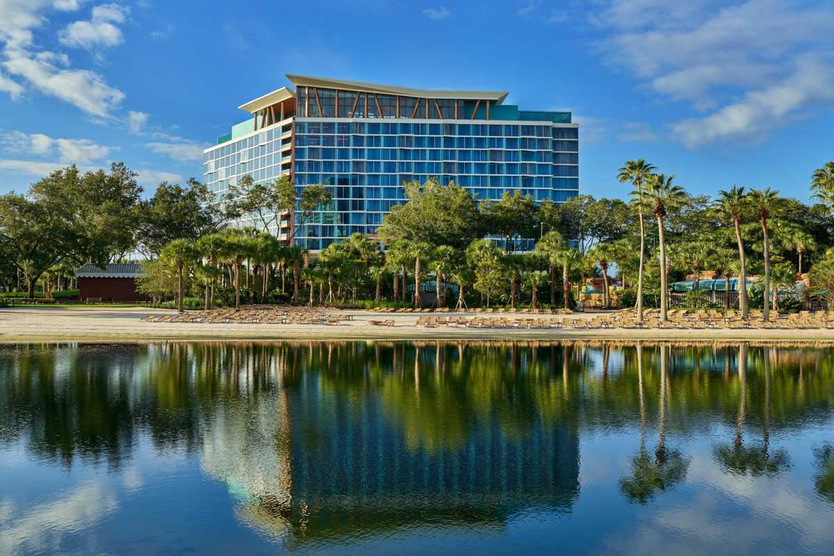 The Swan Reserve Hotel at Walt Disney World features striking blue windows, swaying palm trees, and a shimmering lake reflecting the vibrant sky—offering guests an elegant and immersive resort experience just steps from the magic of Disney’s theme parks.