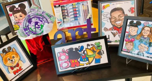 A vibrant display of caricature drawings and art supplies set out on a table, highlighted by a bright purple "art" sign at the center—an inviting stop for Disney Springs guests searching for one-of-a-kind holiday gifts.