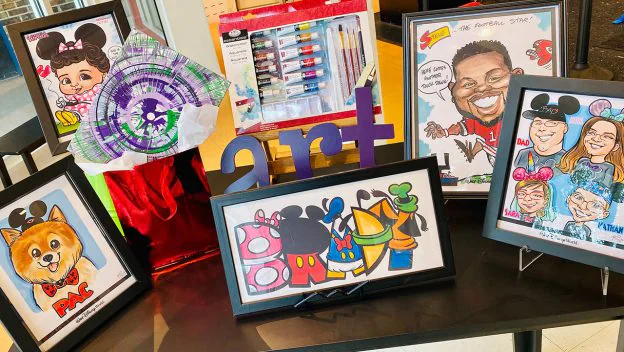 A vibrant display of caricature drawings and art supplies set out on a table, highlighted by a bright purple "art" sign at the center—an inviting stop for Disney Springs guests searching for one-of-a-kind holiday gifts.