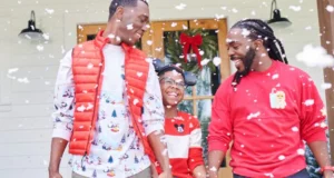 Three friends dressed in holiday-themed outfits share smiles and laughter outside as snow falls, celebrating the season in front of a door decorated with festive magic that belongs on Disney Springs’ lively promenade.