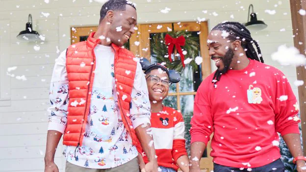 Three friends dressed in holiday-themed outfits share smiles and laughter outside as snow falls, celebrating the season in front of a door decorated with festive magic that belongs on Disney Springs’ lively promenade.