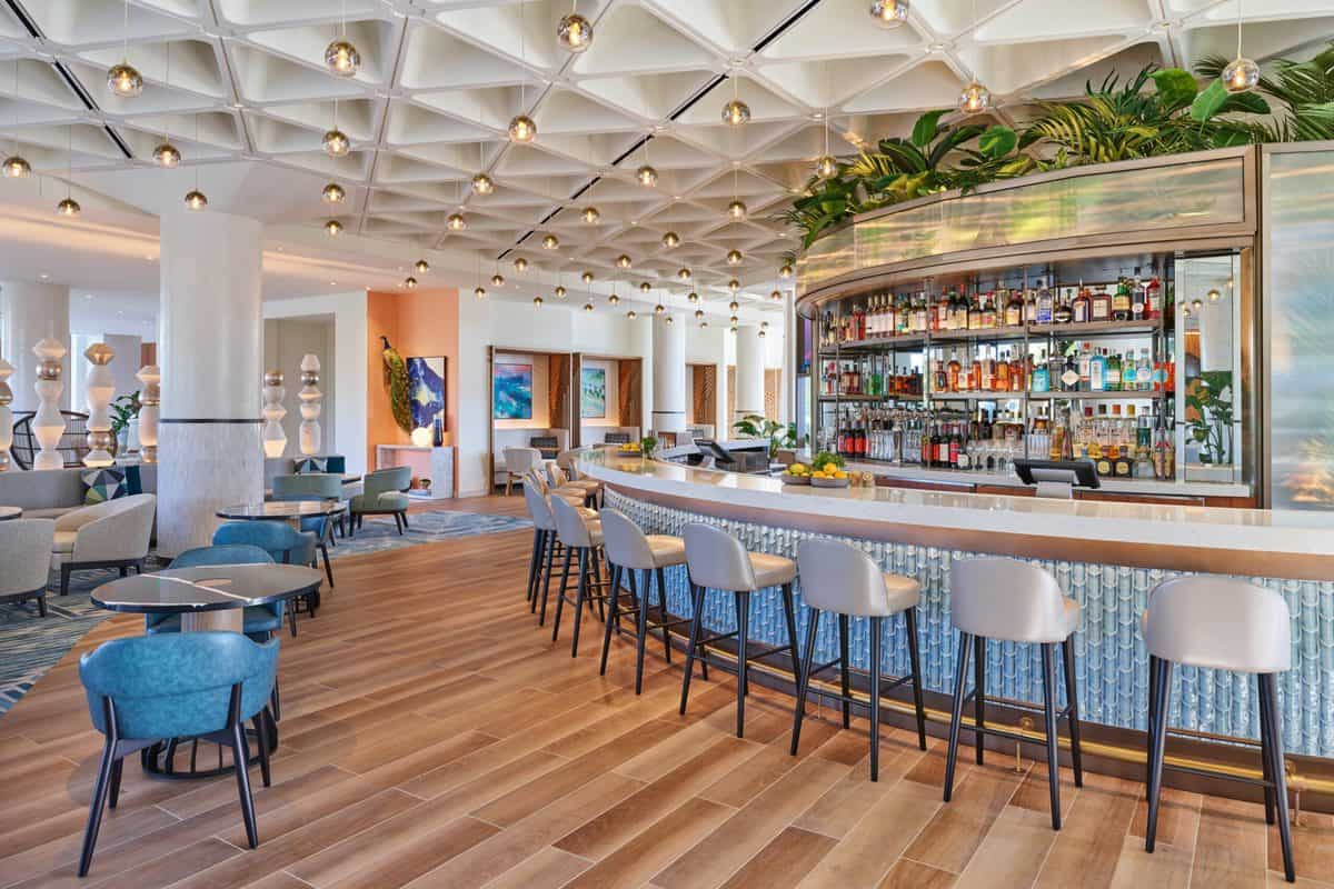 Chic hotel bar at the Swan Reserve Hotel near Walt Disney World, featuring sleek high stools, illuminated liquor shelves, lush greenery above, and modern lounge seating—an inviting spot for park guests to unwind after a day of Disney excitement.