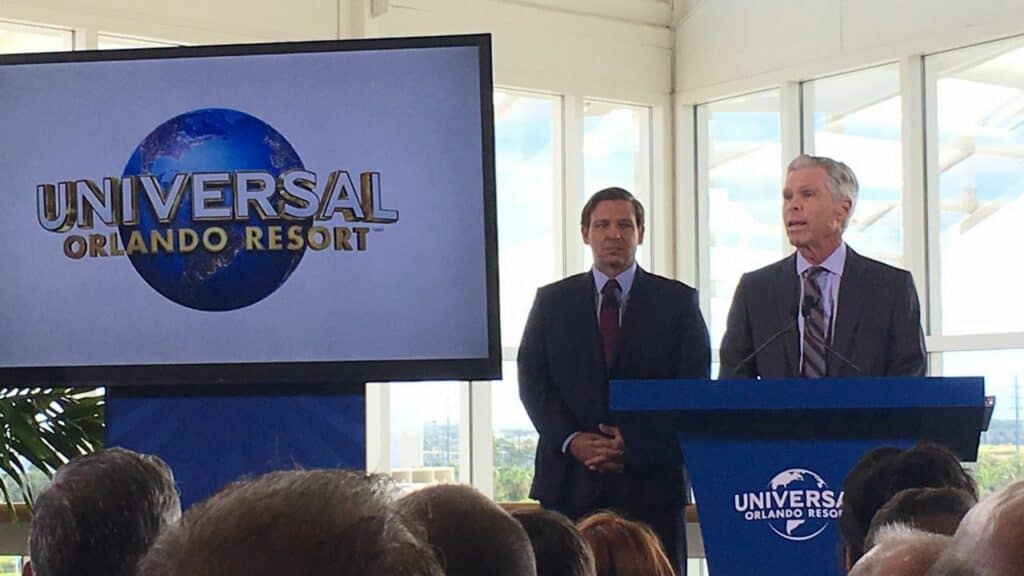 Universal Orlando Resort CEO Tom Williams stands at a podium bearing the Universal logo, making his retirement announcement alongside another executive, with the iconic Universal Orlando Resort branding displayed in the background.