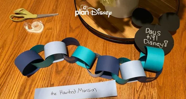 A colorful paper countdown chain, scissors, tape, and a cheerful "Days 'til Disney!" sign rest on a wooden table—essential supplies for Disney fans eagerly anticipating their next magical trip to the parks.
