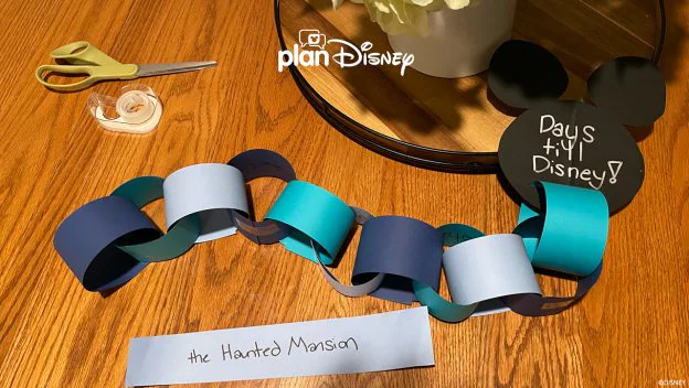 A colorful paper countdown chain, scissors, tape, and a cheerful "Days 'til Disney!" sign rest on a wooden table—essential supplies for Disney fans eagerly anticipating their next magical trip to the parks.