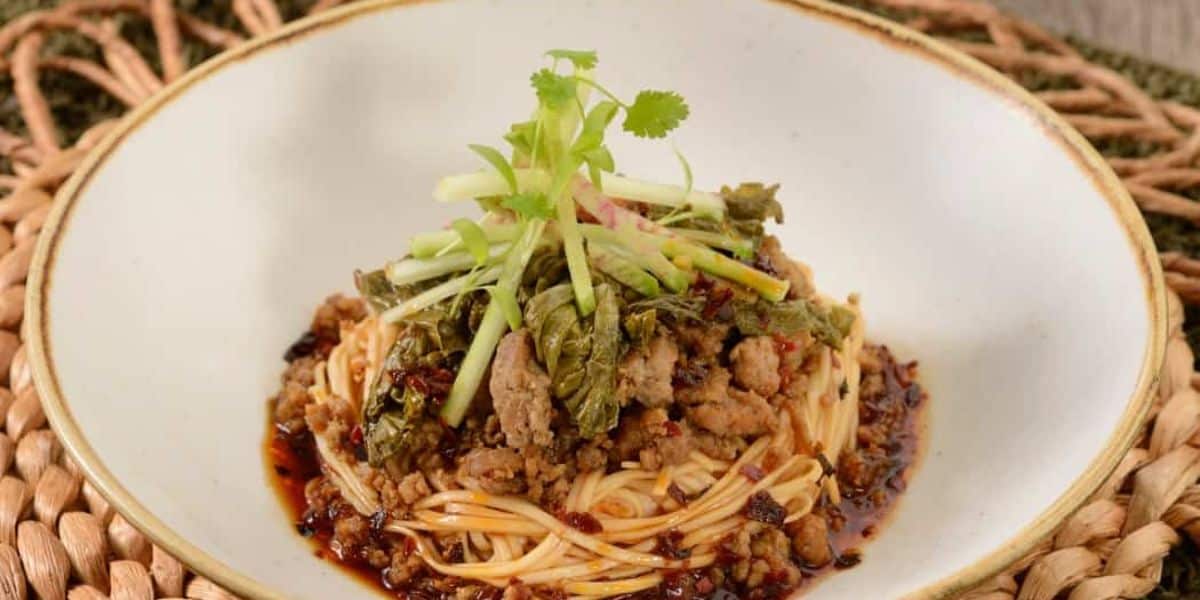 A bowl of noodles topped with minced meat, green vegetables, and garnished with cilantro. The dish is surrounded by a spicy-looking sauce, served on a white plate with a brown rim, placed on a woven mat.