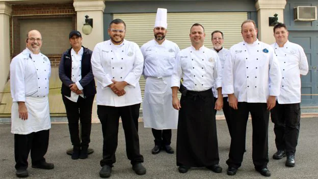 Seven chefs dressed in crisp white uniforms and a team member in a cap gather outside, beaming as they come together for the Coalition for the Homeless’s Thanksgiving outreach—sharing the same camaraderie and joy found among entertainment cast members rallying for a special event.