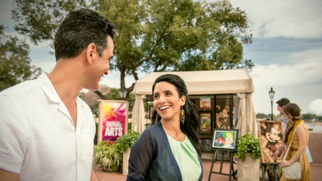 Guests enjoy strolling past colorful art booths at EPCOT International Festival of the Arts, smiling and soaking up the sunshine.