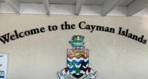 The “Welcome to the Cayman Islands” sign greets visitors like an attraction entrance, featuring a vibrant island crest and inspiring motto, all set on a sunny beige wall beneath a bright white overhang—capturing the inviting spirit of an island adventure destination.