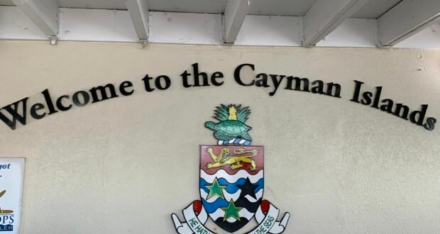 The “Welcome to the Cayman Islands” sign greets visitors like an attraction entrance, featuring a vibrant island crest and inspiring motto, all set on a sunny beige wall beneath a bright white overhang—capturing the inviting spirit of an island adventure destination.
