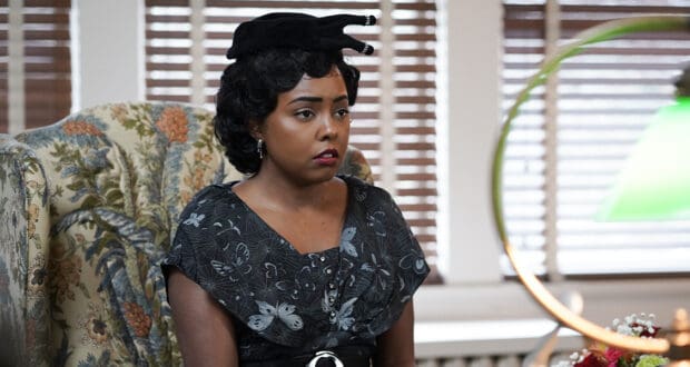 A stylish guest at a classic 2022 Television Edition event, dressed in a vintage black dress and chic hat, relaxes on a floral chair by the window blinds, evoking the glamour and nostalgia found in entertainment park retro-themed lounges.