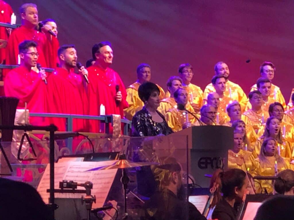 A vibrant choir in red and yellow EPCOT robes performs onstage behind a guest speaker at the podium, with festival musicians in front, filling the theater with the joyful holiday spirit of EPCOT International Festival of the Holidays.