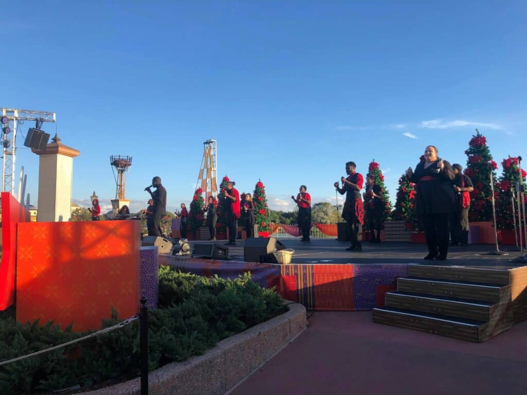 A vibrant choir dressed in red and black lights up the EPCOT International Festival stage, surrounded by sparkling Christmas trees and festive decorations, filling the air with classic holiday music for delighted festival guests.