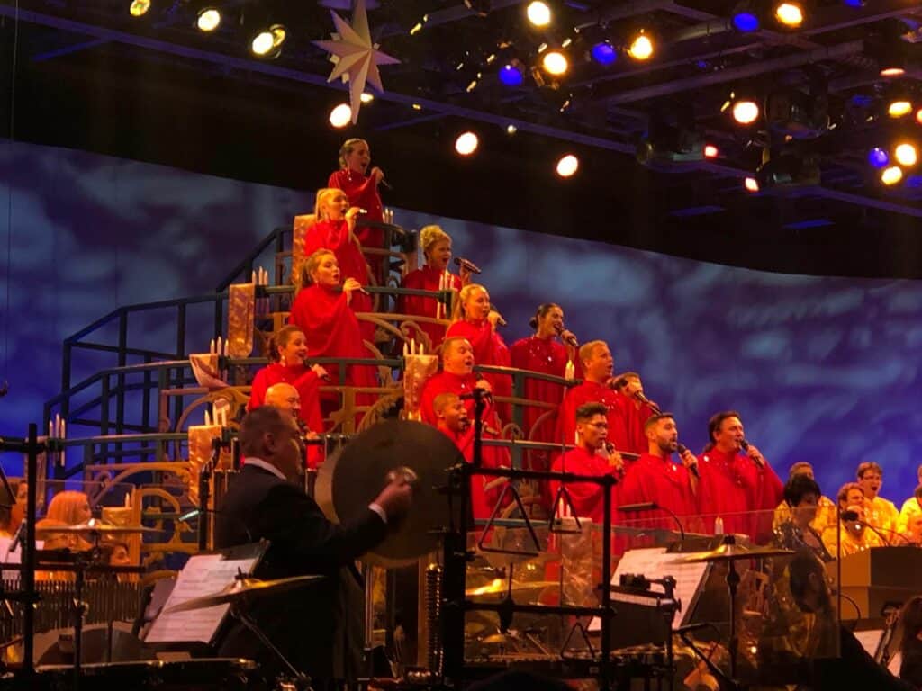 A vibrant choir dressed in red robes performs on tiered risers at EPCOT, their harmonies filling the pavilion with joyful holiday music while musicians play festive tunes in front of them—creating an unforgettable holiday entertainment experience at the EPCOT International Festival.