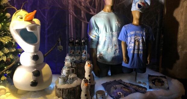 Olaf and other Frozen merchandise on display at EPCOT’s Festival of the Holidays, with themed mugs, shirts, and water bottles arranged on a wintry, snow-dusted table.