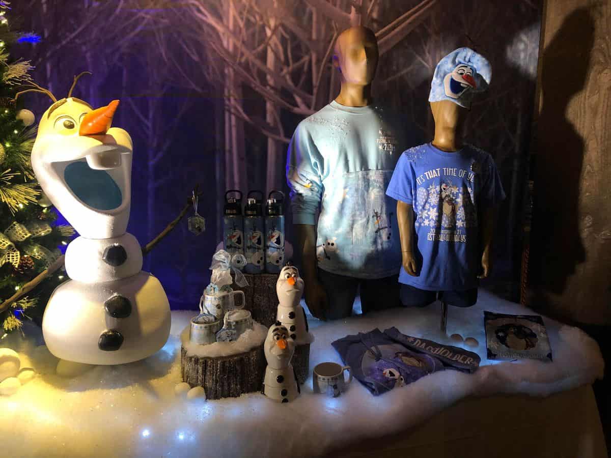 Olaf and other Frozen merchandise on display at EPCOT’s Festival of the Holidays, with themed mugs, shirts, and water bottles arranged on a wintry, snow-dusted table.