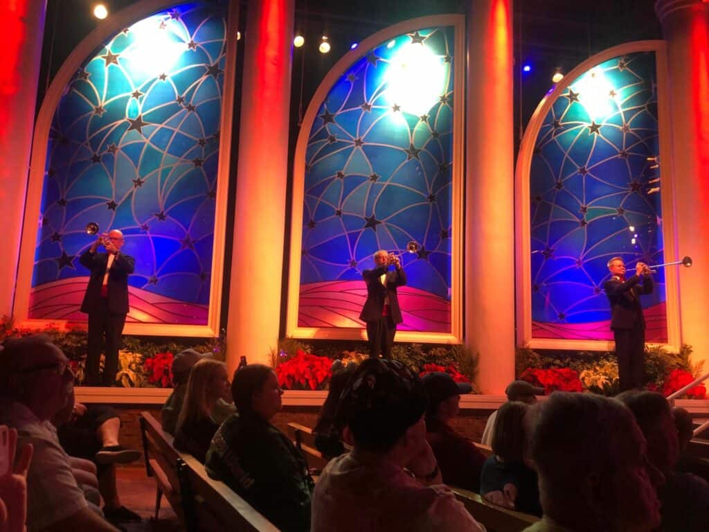 Trumpet players light up the stage at EPCOT International Festival, performing in front of vibrant blue arches and red lighting as festival guests enjoy the lively show from their seats.
