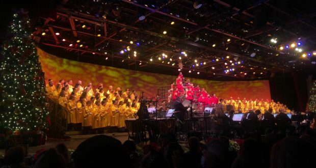 A choir in shimmering gold robes performs holiday music on stage at EPCOT, surrounded by a sparkling Christmas tree and festive lights, capturing the magical spirit of the International Festival of the Holidays.