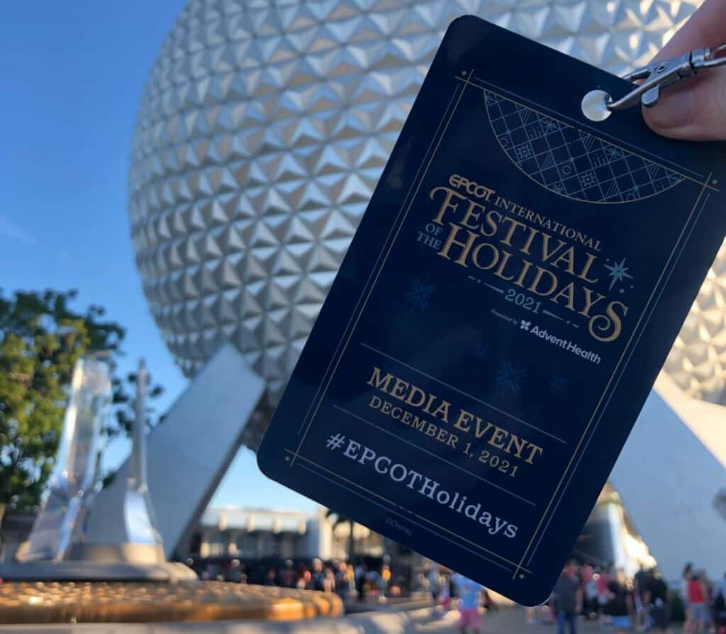 An EPCOT International Festival of the Holidays media pass displayed in front of the iconic Spaceship Earth, highlighting the festive atmosphere and lively holiday entertainment throughout EPCOT.