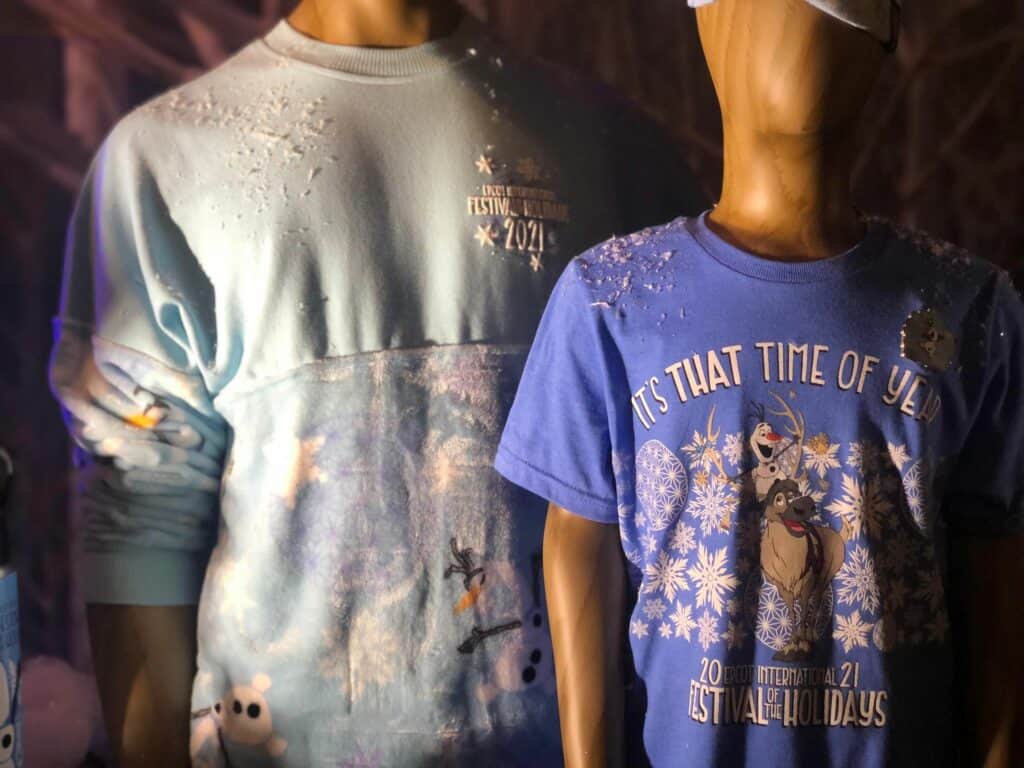 At EPCOT's Festival of the Holidays, two mannequins showcase blue Disney holiday shirts featuring Olaf and Sven surrounded by playful patches of faux snow, highlighting the exclusive winter merchandise perfect for fans celebrating the magic of the season.