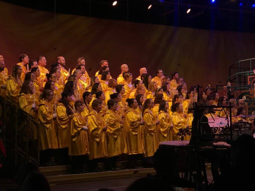 The holiday choir at Disneyland Resort, dressed in bright yellow robes, sings festive tunes on stage beneath warm lights. The performers face the conductor and musicians, filling the park with magical holiday cheer and creating unforgettable entertainment for guests.