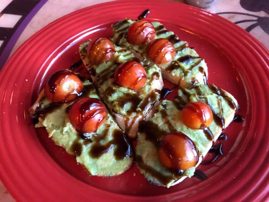 Avocado toast topped with cherry tomatoes and a drizzle of balsamic glaze, served on a vibrant red plate—one of the whimsical breakfast choices available at Goofy's Kitchen in Disneyland, where guests enjoy inventive dishes inspired by Disney magic.