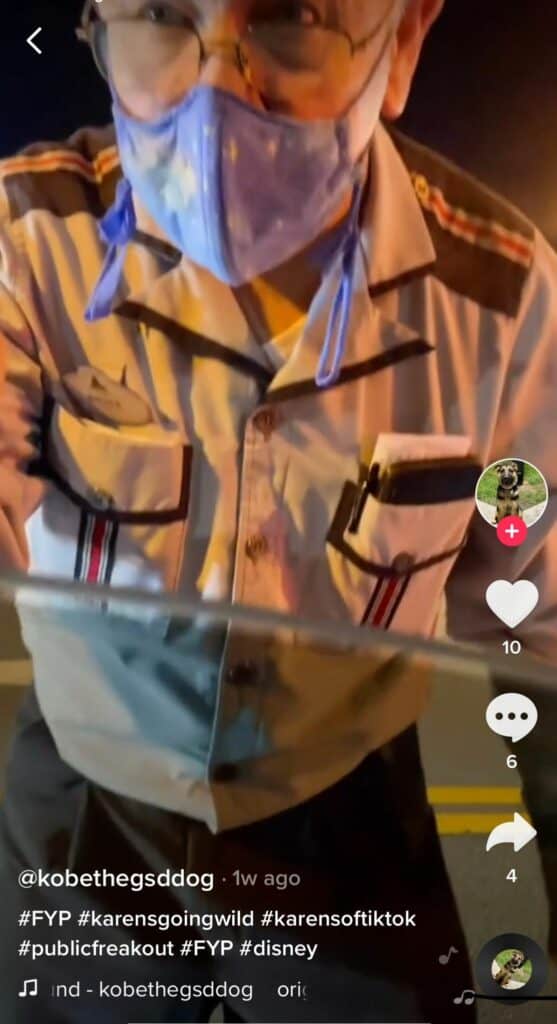 A Disney cast member in themed uniform and a blue face mask leans toward the camera under nighttime park lights, with a TikTok interface overlay on the screen.
