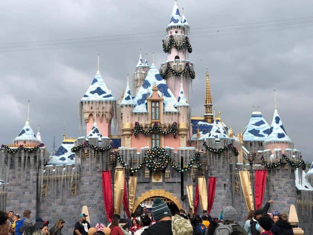 Guests flock to Sleeping Beauty Castle at Disneyland, decked out in sparkling snow and shimmering holiday garlands, as a magical winter scene unfolds beneath a cloudy California sky.