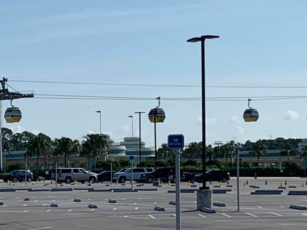 In a mostly empty Disney World parking lot with scattered cars, iconic Skyliner gondolas gliding overhead, and palm trees swaying in the background, a dramatic confrontation unfolds between a Disney Cast Member and a Guest.