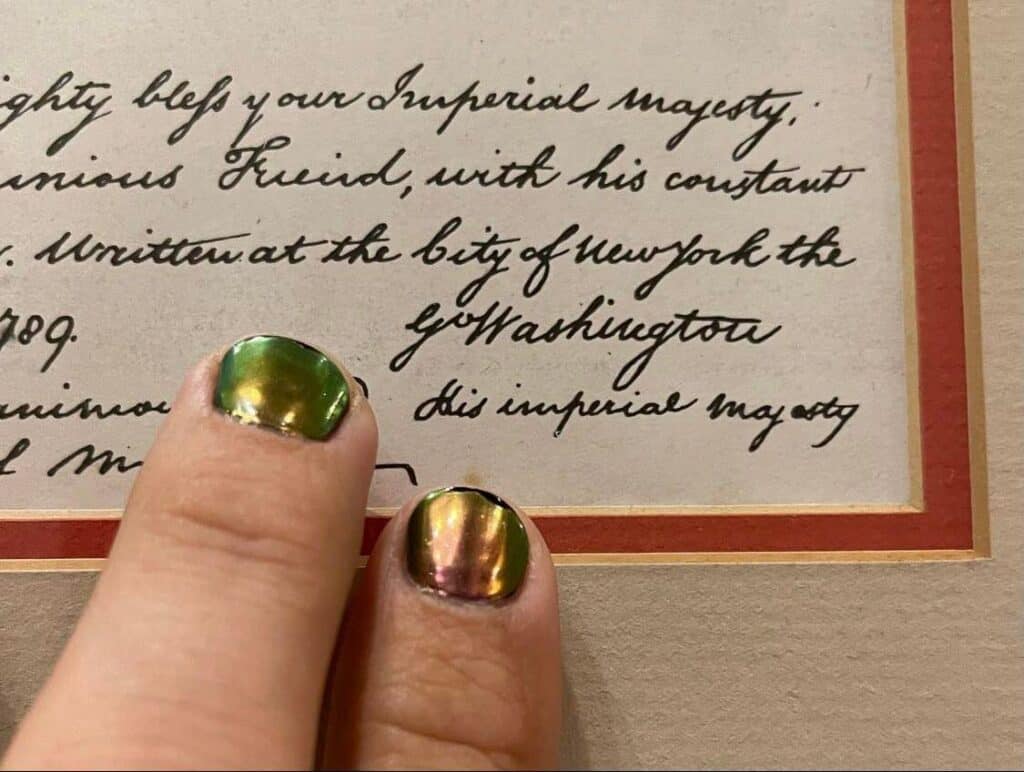 Fingers with shimmering green polish highlight George Washington’s signature on an antique document, evoking the immersive storytelling and design details found in EPCOT’s Morocco Pavilion, as featured by Imagineer Zach Riddley.
