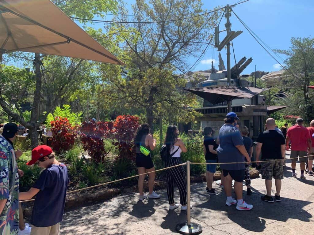 Guests line up outside Space Mountain at Magic Kingdom on a bright, sunny day, waiting near lush landscaping and the iconic spired roof with antennas. Tempers flare among some visitors as they voice their frustration over the ride’s recent breakdown.