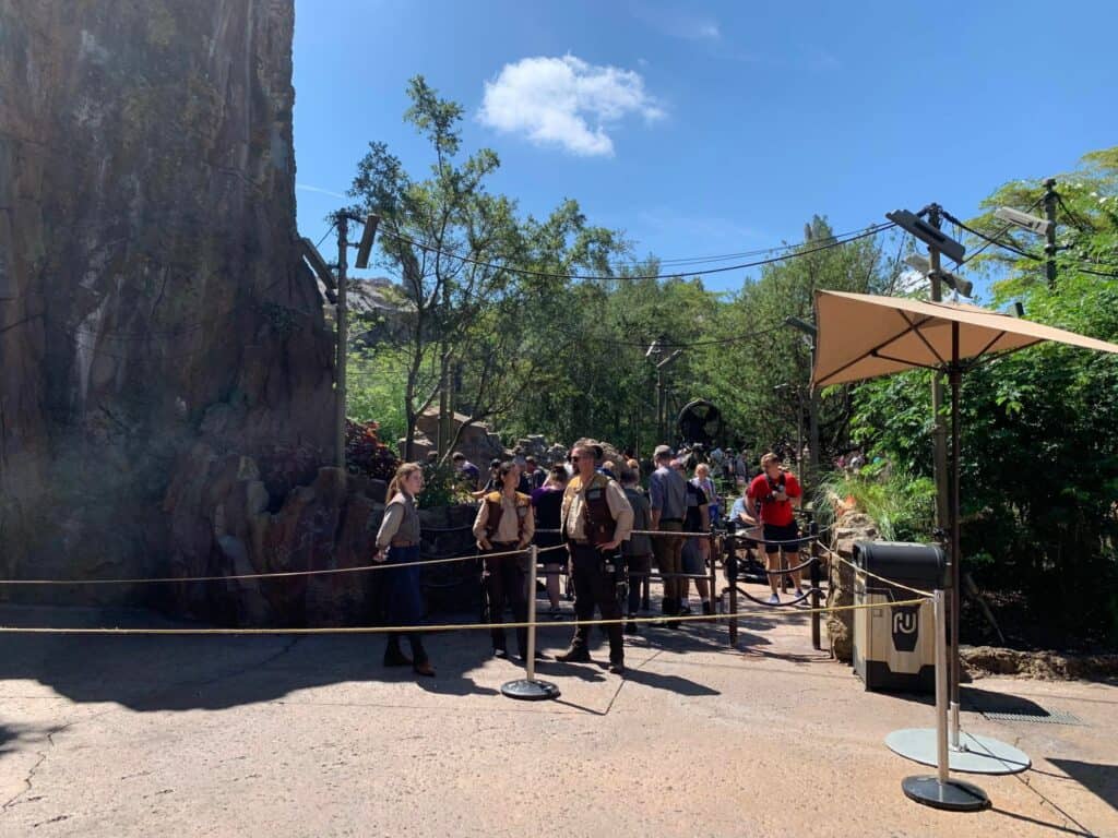 Guests line up outside in Adventureland, surrounded by lush landscaping and rocky outcrops under the sun, as staff members assist near a shaded umbrella; frustrated visitors can be seen voicing their disappointment following an unexpected Jungle Cruise ride closure.