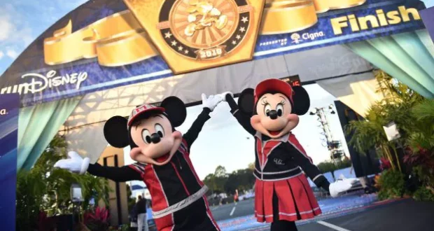 Mickey and Minnie Mouse, dressed in dynamic racing outfits, joyfully celebrate at the finish line of a runDisney race beneath a grand arch, bringing the electric atmosphere of Disney’s 2023 race season to life for fans and guests.
