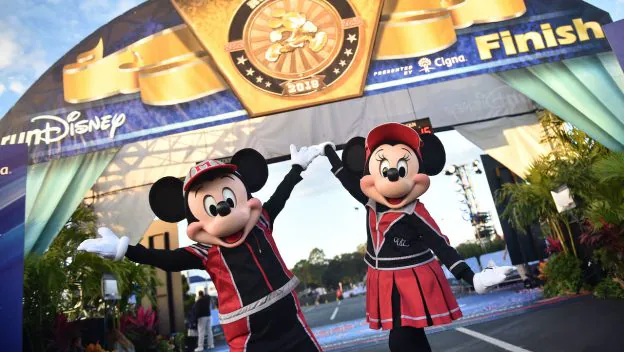 Mickey and Minnie Mouse, dressed in dynamic racing outfits, joyfully celebrate at the finish line of a runDisney race beneath a grand arch, bringing the electric atmosphere of Disney’s 2023 race season to life for fans and guests.