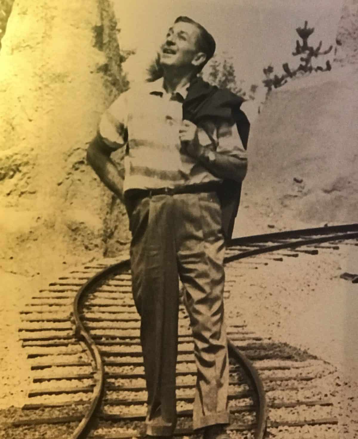 Walt Disney, dressed in nostalgic attire, smiles cheerfully while standing on a winding railroad track beside rugged rocks, capturing the joyful birthday spirit that echoes throughout Disneyland.