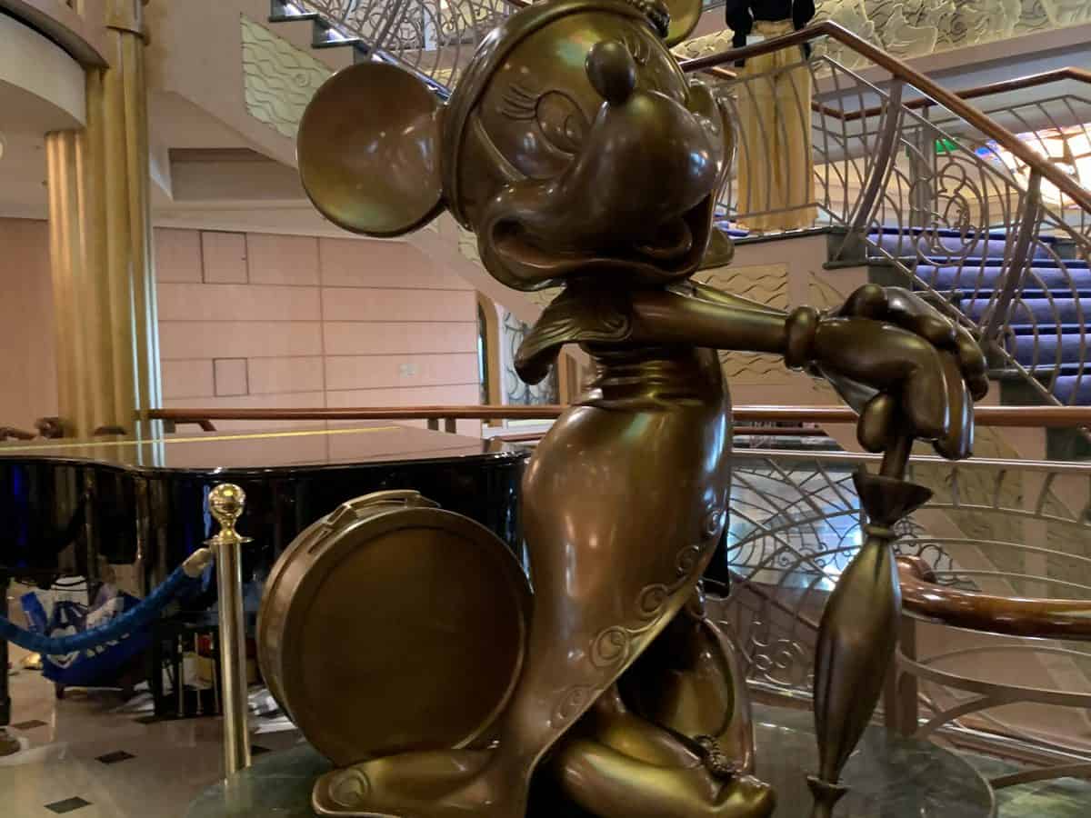 Bronze Minnie Mouse statue holding an umbrella, elegantly showcased indoors near a piano and staircase—a charming Disney Cruise Line icon captivating guests as they explore the ship and its magical destinations.