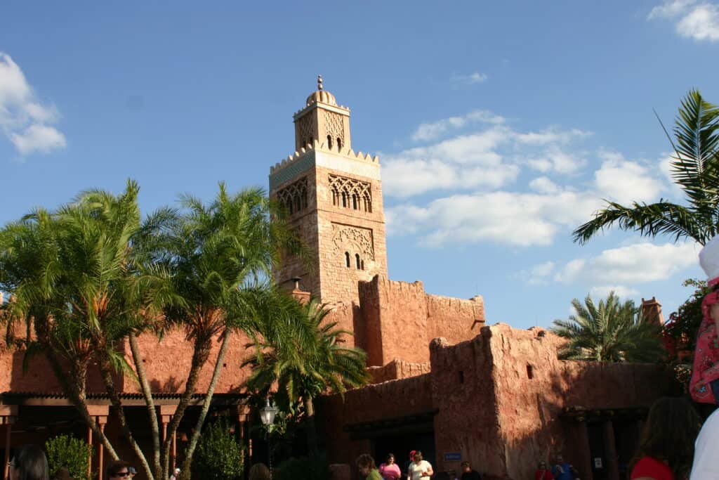 The iconic Moroccan-style tower rises above swaying palm trees under a clear blue sky in EPCOT’s lively Morocco Pavilion, where guests stroll through immersive spaces crafted with Disney Imagineering magic under Zach Riddley’s creative vision.
