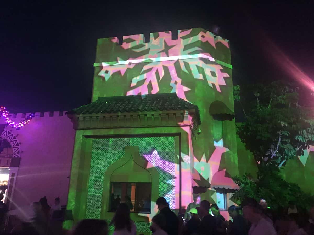 The exterior of a Disney World building glows at night with dazzling green and pink snowflake-shaped light projections, setting a magical, party-like mood that’s perfect for New Year’s Eve festivities in the park.
